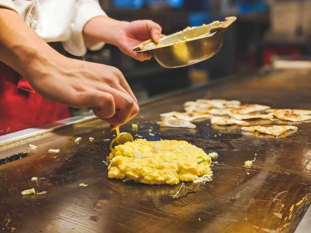 Chef preparing teppanyaki on a hot grill with dumplings cooking in the background.