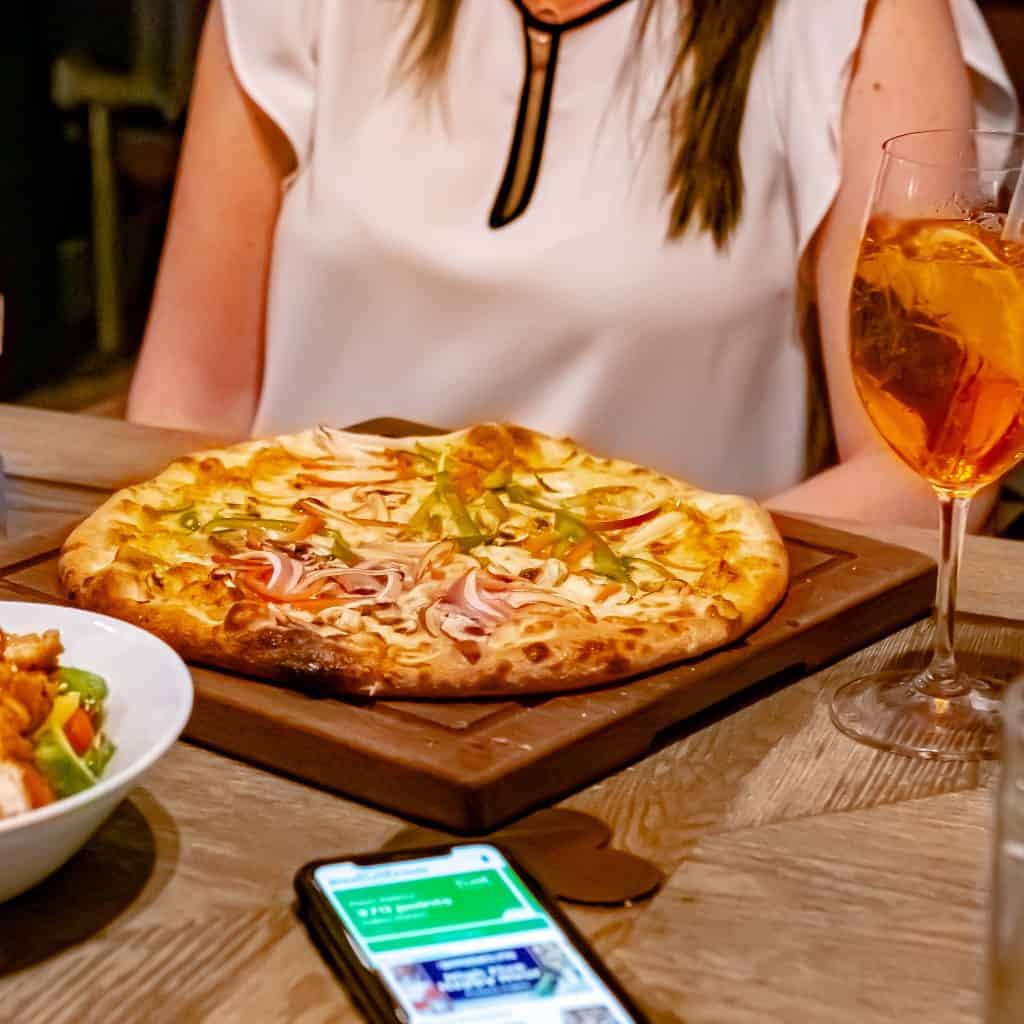 Woman enjoying pizza at a restaurant while using the Bite Club rewards app on her smartphone.